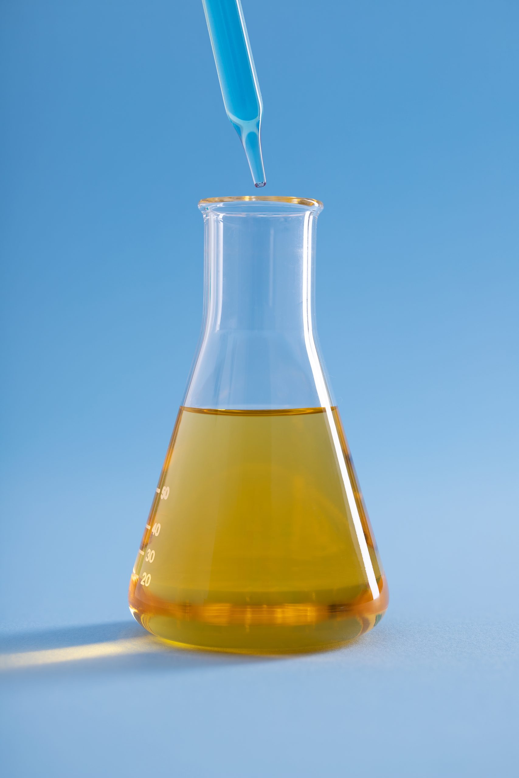 Vertical closeup of dropper with blue liquid over Erlenmeyer flask yellow liquid on blue background